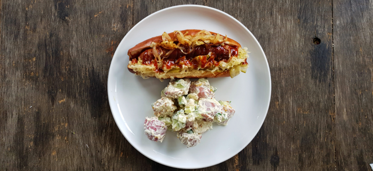 Ananda hot dog with dill pickle kraut, and BBQ sauce on a pretzel bun with creamy potato celery salad